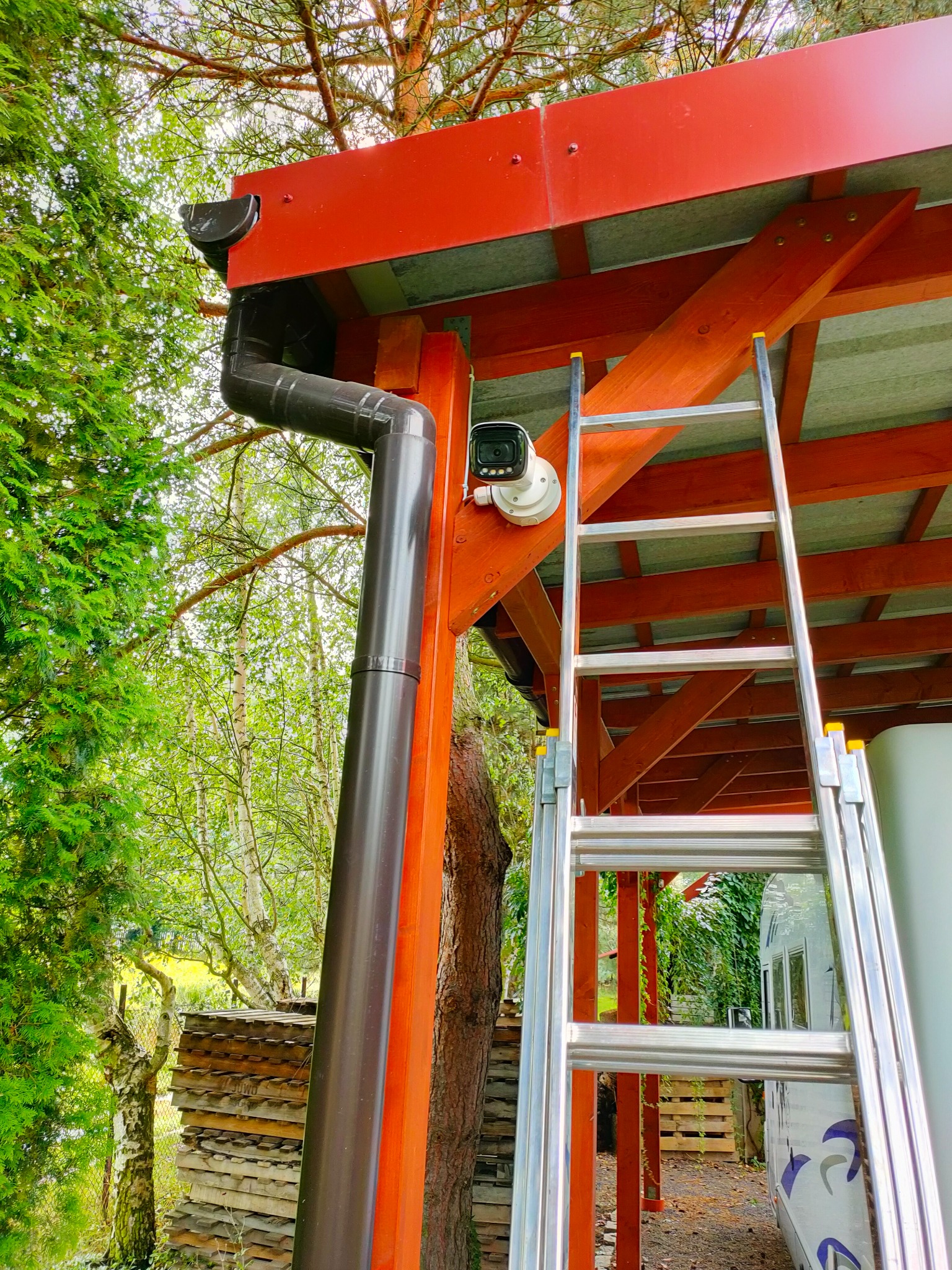 Kamera monitoringu zamontowana na drewnianej konstrukcji z rynną, widoczna drabina i zieleń drzew w tle.