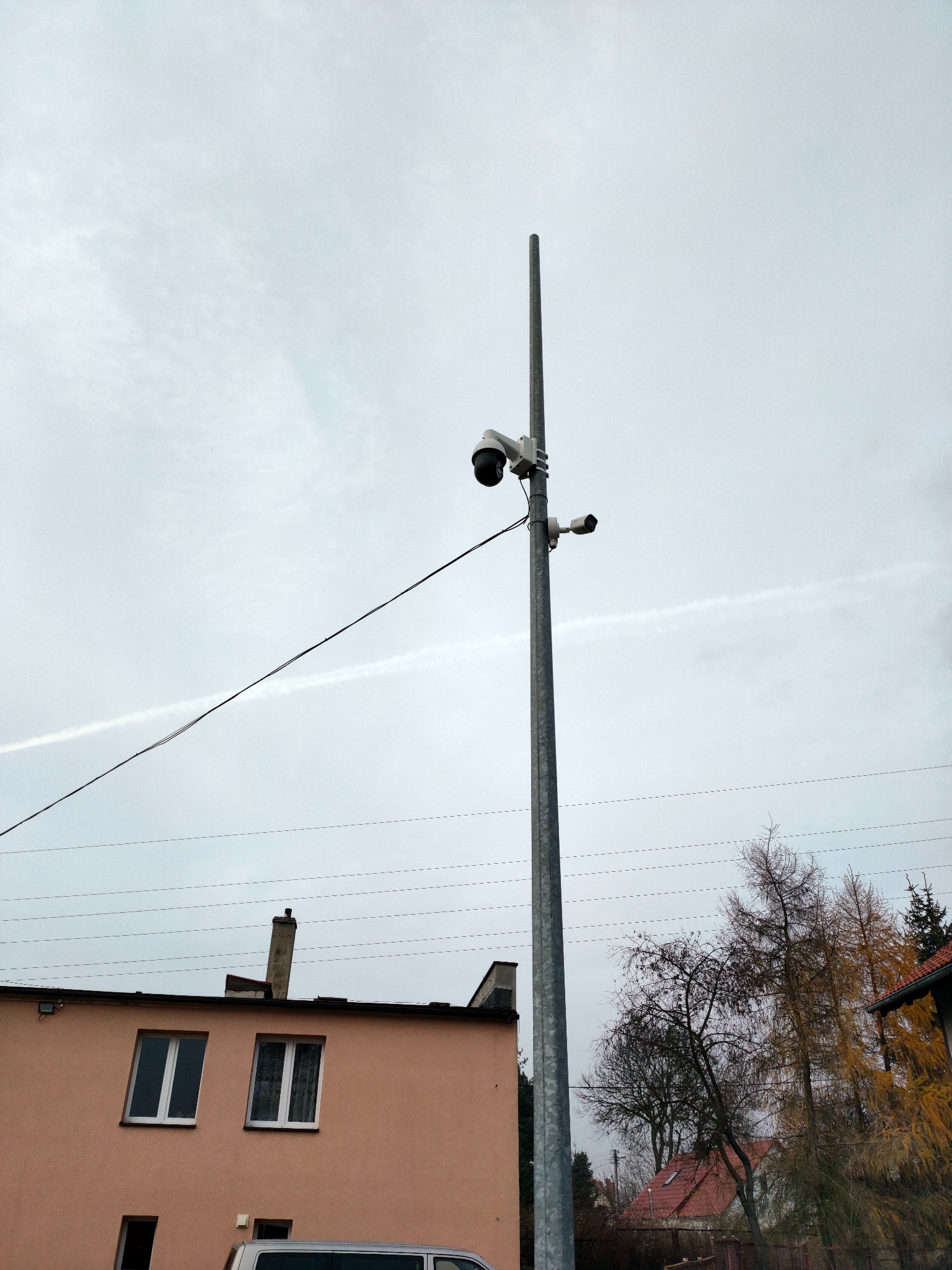 Dwie kamery monitoringu zamontowane na wysokim, metalowym słupie na tle pochmurnego nieba i budynku mieszkalnego z widocznymi liniami energetycznymi.