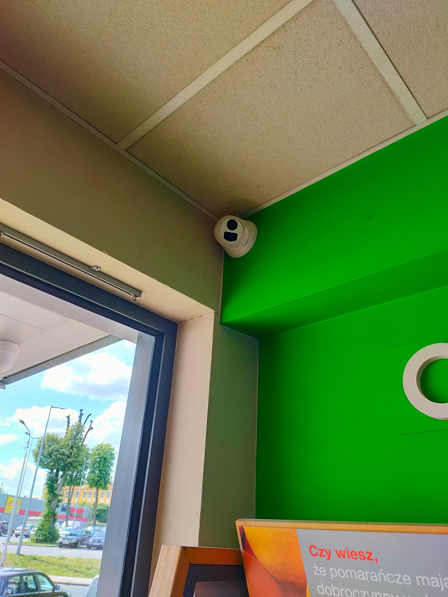 Kamera kopułkowa zamontowana w rogu pomieszczenia z jaskrawozielonymi ścianami, widok z dołu na sufit i fragment okna z widokiem na ulicę.