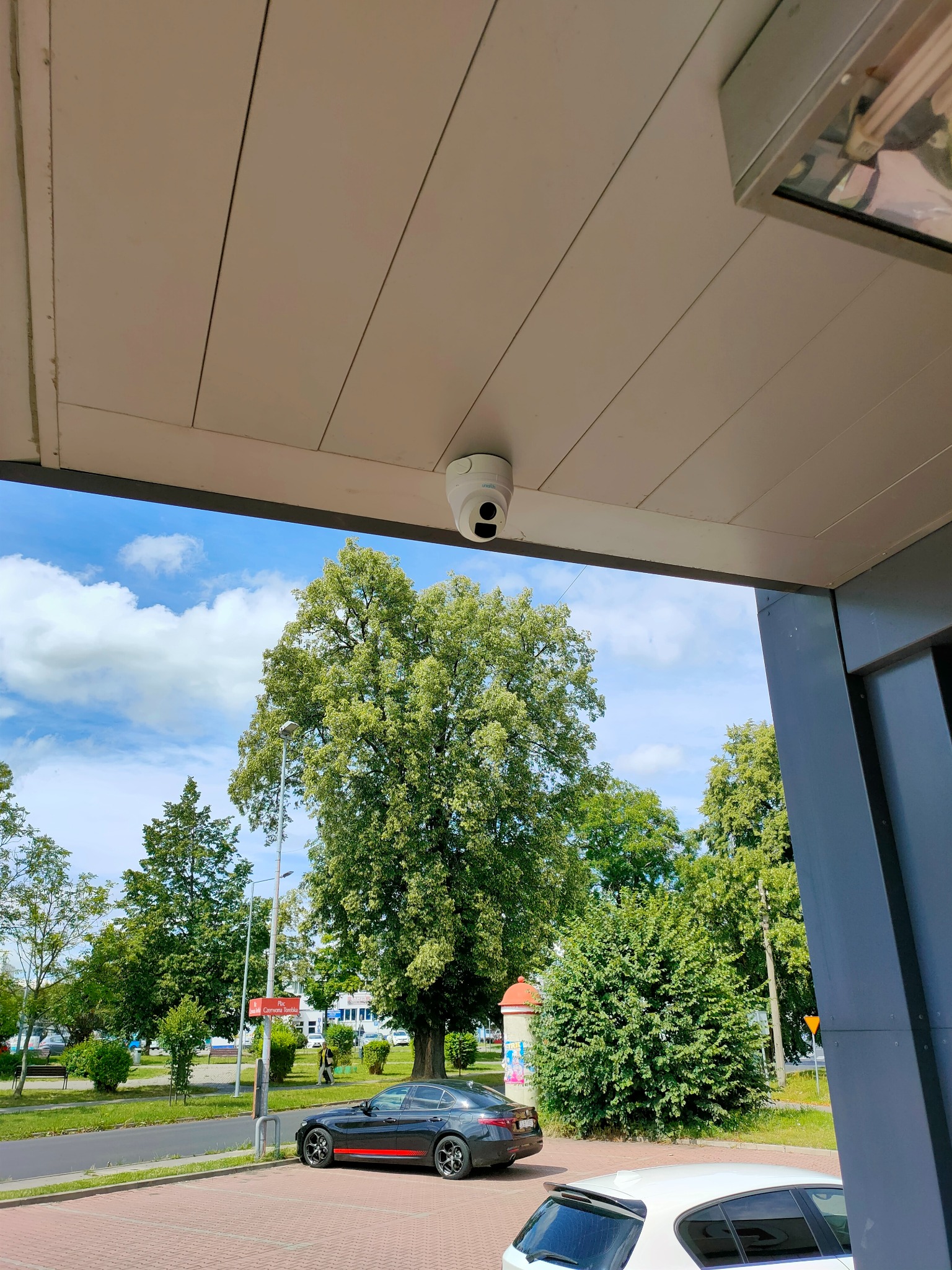 Kopułkowa kamera monitoringu zamontowana pod zadaszeniem, widok na drzewa i zaparkowane samochody w tle.