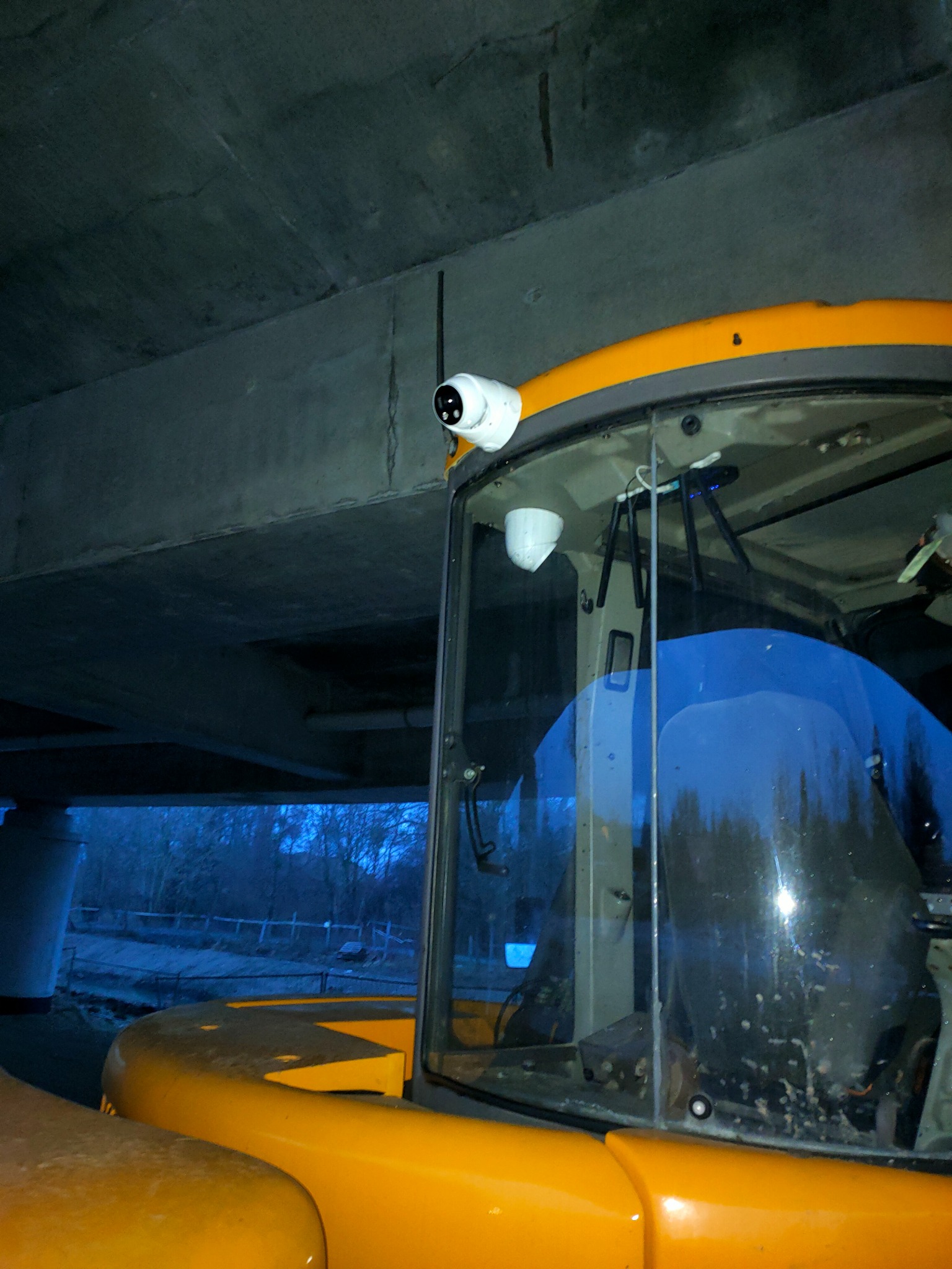 Dwie białe kamery przemysłowe zamontowane na dachu kabiny żółtej maszyny budowlanej pod betonowym wiaduktem, widoczne okablowanie i uchwyty montażowe.