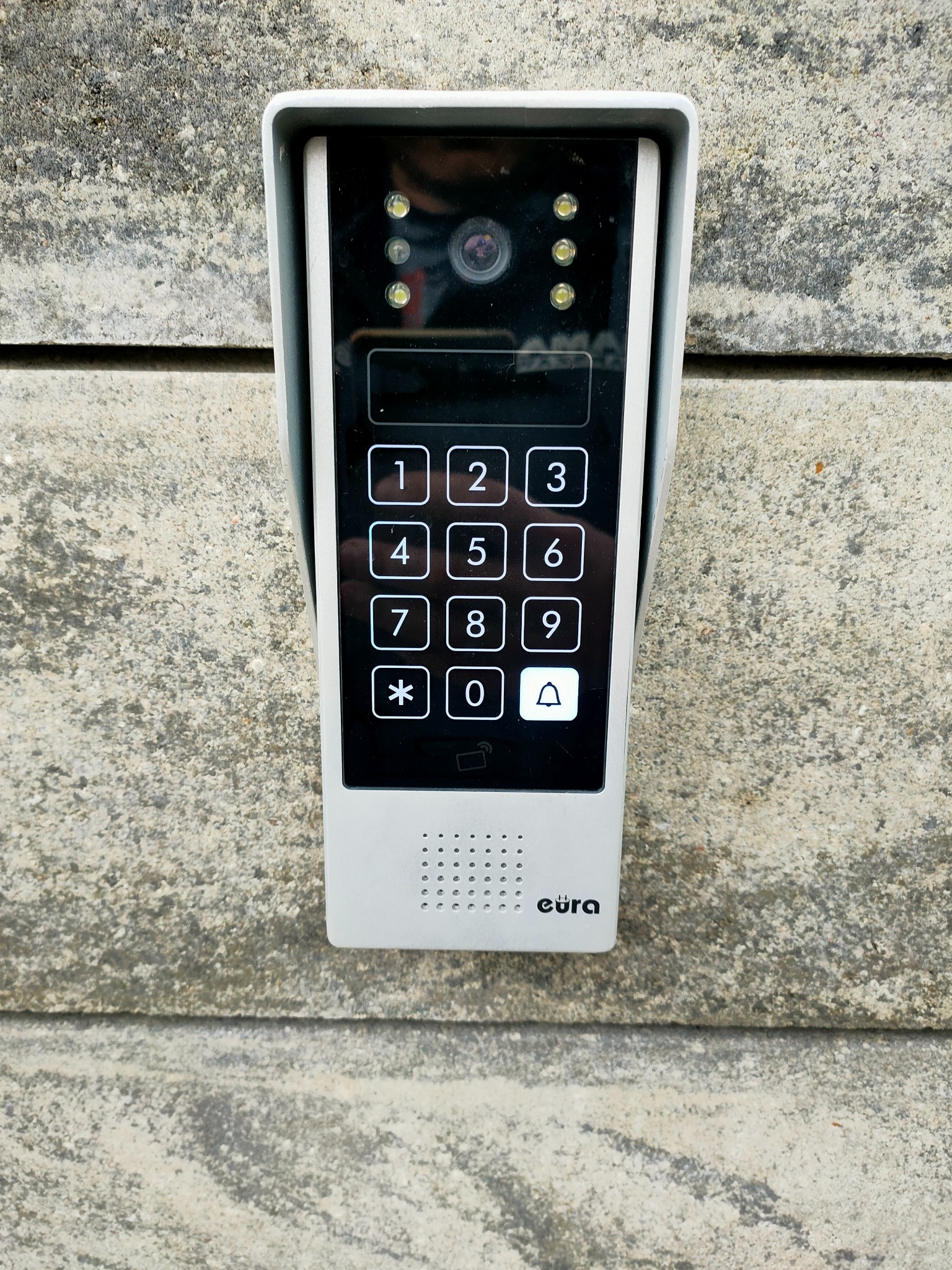 Panel zewnętrzny wideodomofonu Eura z klawiaturą numeryczną i kamerą, zamontowany na szarej, betonowej ścianie.