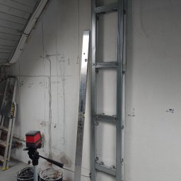 Radosław Butryński Korso-Remonty - Szkielet konstrukcji metalowej przy ścianie z odręcznymi notatkami i poziomica w trakcie prac wykończeniowych we Wrocławiu; widoczny laser krzyżowy, drabina i wiadro z zaprawą.