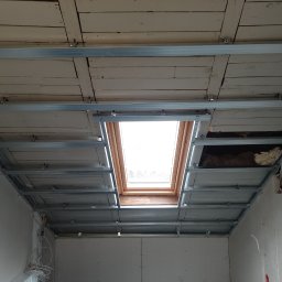 Radosław Butryński Korso-Remonty - Montaż stelaża pod płytę karton gips na suficie z oknem dachowym, widoczne stare deski sufitowe i izolacja.