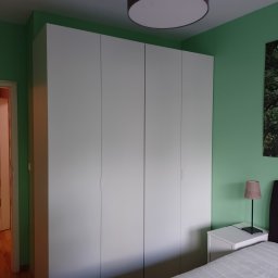 Radosław Butryński Korso-Remonty - Wnętrze sypialni z zielonymi ścianami, białym, czterodrzwiowym wbudowanym szafą, lampką nocną na białej szafce i częścią łóżka z materacem.