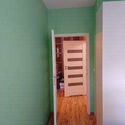 Radosław Butryński Korso-Remonty - Ściana pomalowana na zielono, widok na otwarte drzwi z panelami i fragment szafy. Na podłodze panele drewnopodobne.