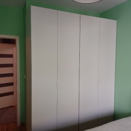 Radosław Butryński Korso-Remonty - Pokój z zielonymi ścianami, białym czterodrzwiowym segmentem szafy i widocznym fragmentem łóżka z zagłówkiem oraz otwartymi drzwiami do innego pomieszczenia.