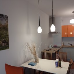 Radosław Butryński Korso-Remonty - Jasne wnętrze mieszkania z pomarańczowymi akcentami, widok na stół z dekoracją z suszonych traw, obraz na ścianie i oświetlenie sufitowe.