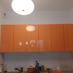 Radosław Butryński Korso-Remonty - Pomarańczowe szafki kuchenne pod sufitem z widocznymi refleksami światła, poniżej zlewozmywak z naczyniami i butelkami.