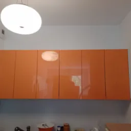 Pomarańczowe szafki kuchenne wiszące z błyszczącymi frontami, oświetlone okrągłą lampą sufitową, widok od dołu.