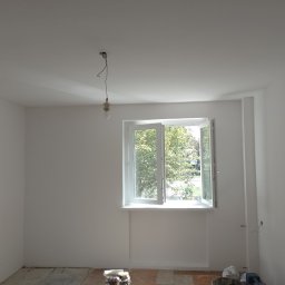 Radosław Butryński Korso-Remonty - Jasne, świeżo pomalowane wnętrze pokoju z otwartym oknem, widoczne narzędzia malarskie na podłodze i wisząca żarówka bez klosza.