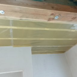 Radosław Butryński Korso-Remonty - Montaż folii na drewnianej konstrukcji sufitu podwieszanego w pomieszczeniu z pomalowanymi ścianami, widoczne śruby mocujące.