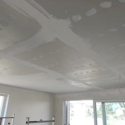 Radosław Butryński Korso-Remonty - Nierówna powierzchnia sufitu z widocznymi łatami i świeżo nałożoną gładzią, przygotowywana do dalszych prac wykończeniowych, z widocznymi przewodami elektrycznymi i fragmentem drabiny w tle.