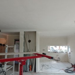 Radosław Butryński Korso-Remonty - Wnętrze w trakcie prac wykończeniowych: gładkie, białe ściany i sufit, widoczny podnośnik do płyt gipsowych, odkurzacz przemysłowy i narzędzia, okno z widokiem na ulicę.