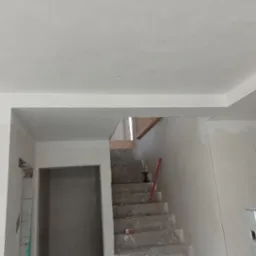 Wnętrze w trakcie prac wykończeniowych: surowe schody betonowe, ściany po nałożeniu gładzi, widoczny poziomica, otwór drzwiowy z widocznymi bloczkami i zapiskami, sufit z gładzią