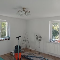 Radosław Butryński Korso-Remonty - Pokój po świeżym malowaniu ścian na biało, widoczne okna, grzejnik, lampa sufitowa, drabina, wentylator, odkurzacz przemysłowy oraz panele podłogowe w opakowaniach.