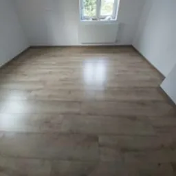 Jasnobrązowe panele podłogowe ułożone w pustym, jasnym pokoju na poddaszu ze skosami i oknem. Widoczny biały grzejnik pod oknem.