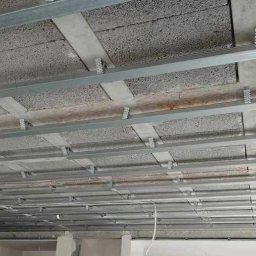 Radosław Butryński Korso-Remonty - Montaż metalowego stelaża sufitu podwieszanego, widoczne profile i elementy mocujące do betonowego stropu, wystające przewody elektryczne.