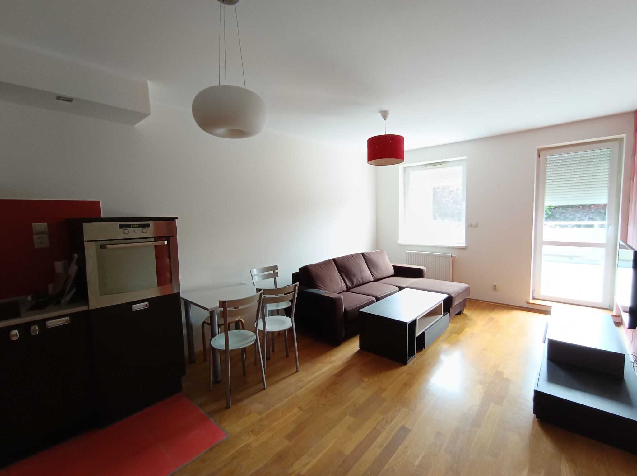 Jasne wnętrze mieszkania z drewnianą podłogą, brązową sofą z szezlongiem, stolikiem kawowym, stołem z krzesłami i zabudową kuchenną w odcieniach czerwieni i czerni, oświetlone dwoma lampami...