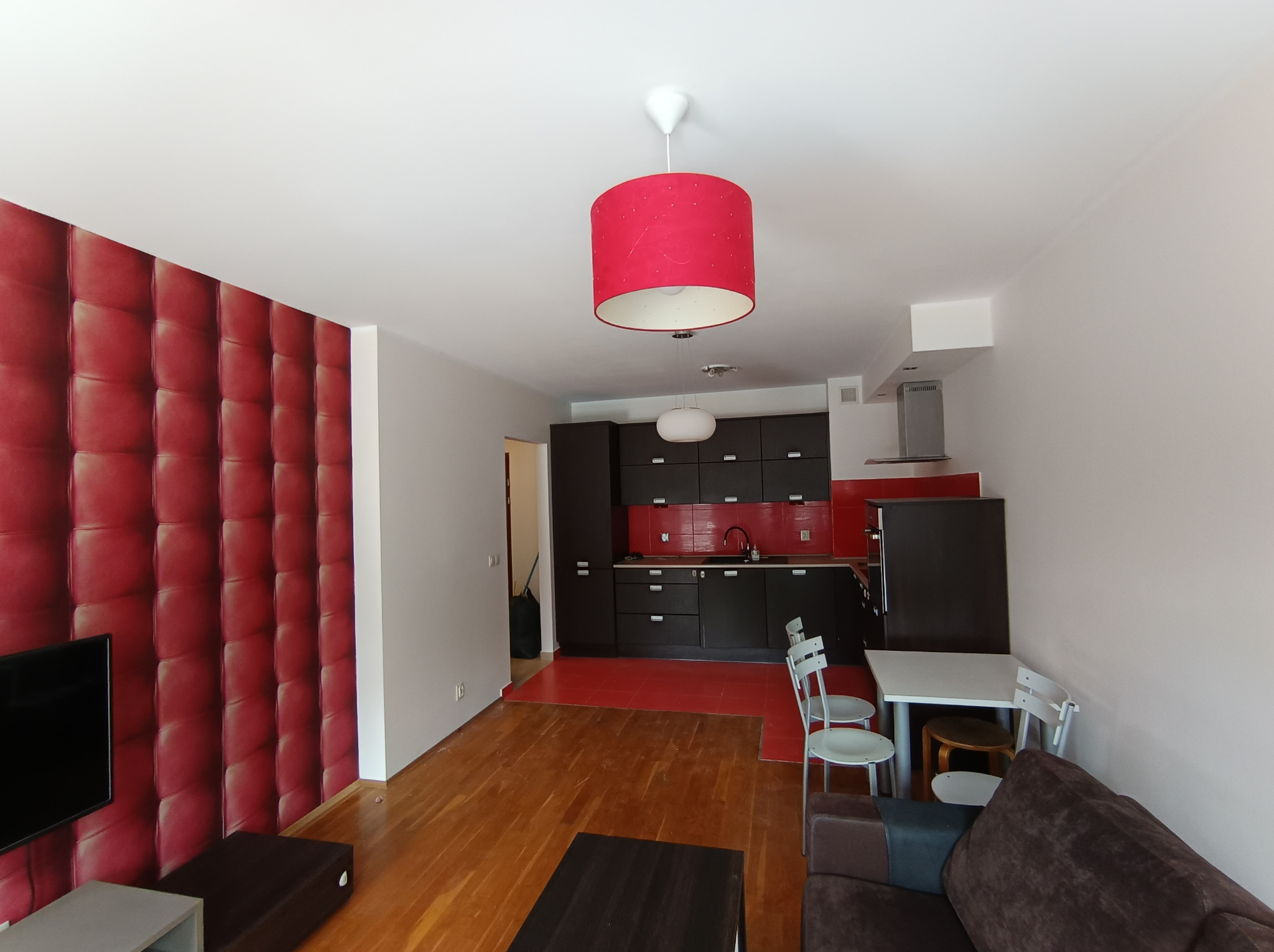 Widok na salon połączony z kuchnią, z dominującą czerwoną ścianą z efektem pikowania, drewnianą podłogą i czerwonymi akcentami w kuchni, oświetlony czerwoną lampą wiszącą z białym wnętrzem.