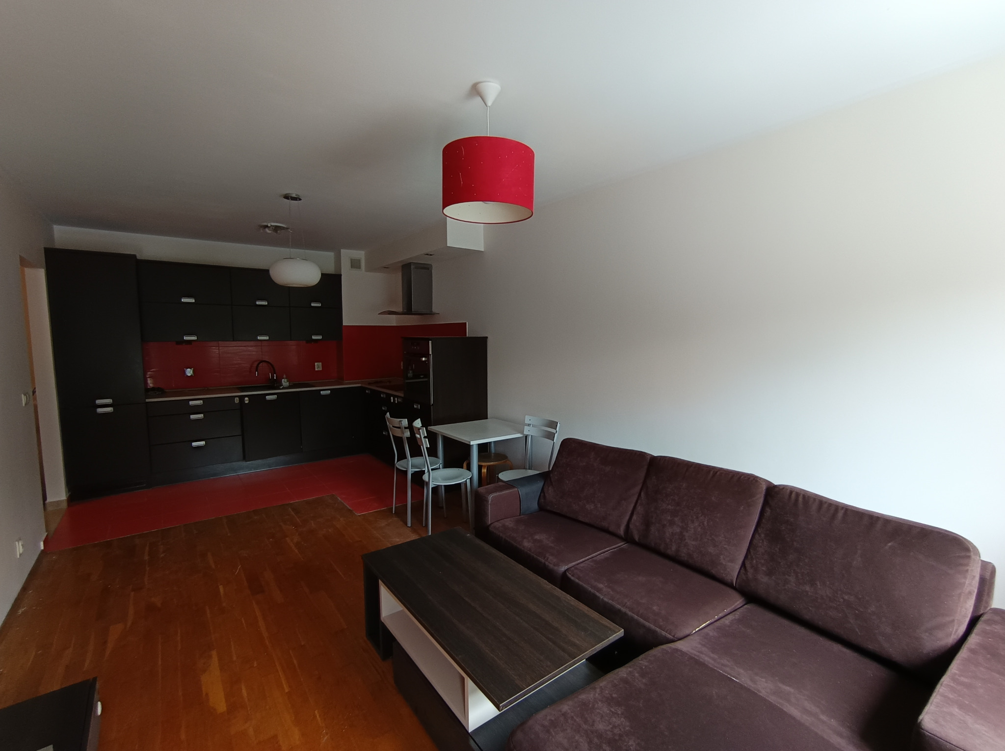 Nowoczesne wnętrze mieszkania z ciemną zabudową kuchenną, czerwonymi akcentami i brązowym narożnikiem, widok z perspektywy podłogi.