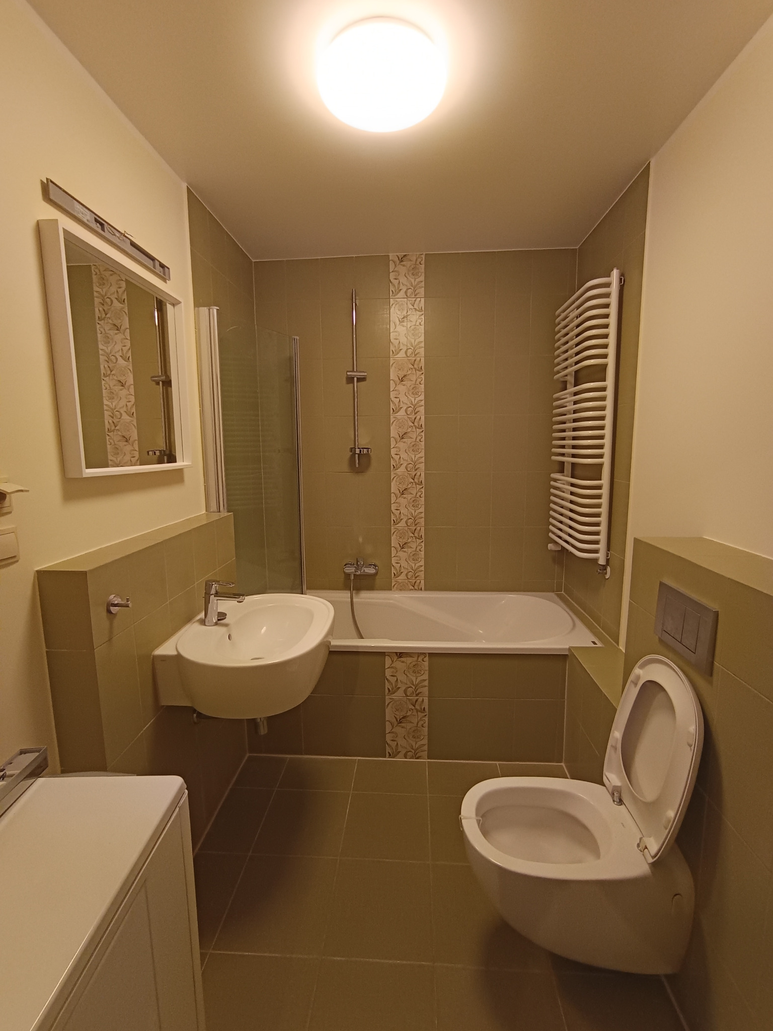 Wykończona łazienka w odcieniach oliwkowej zieleni z dekoracyjnymi płytkami, białą armaturą i grzejnikiem drabinkowym.