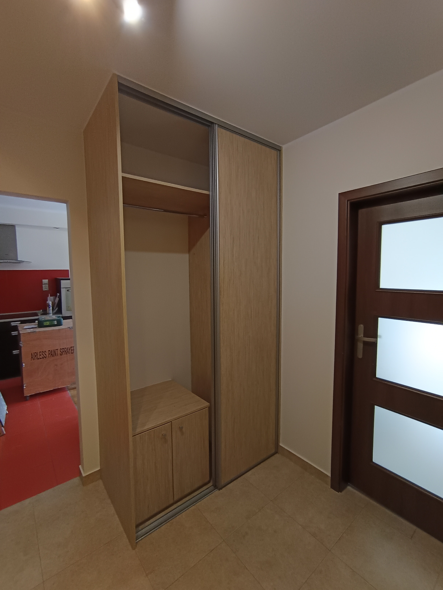 Wnętrze mieszkania z zamontowaną szafą wnękową z przesuwnymi drzwiami o drewnopodobnej fakturze, widoczny fragment kuchni z czerwonymi panelami i karton z napisem 'AIRLESS PAINT SPRAYER'.
