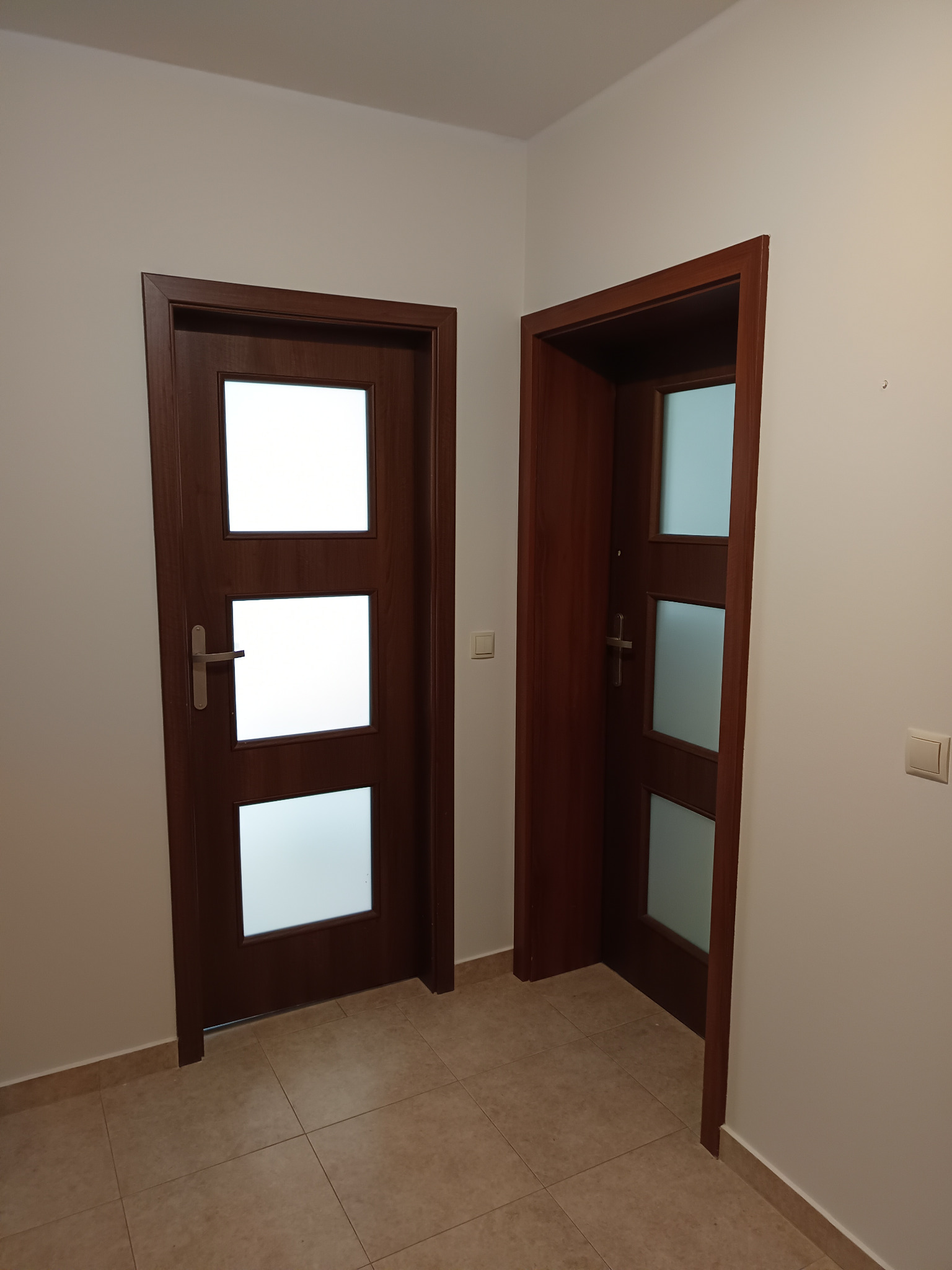 Dwie zamontowane, ciemnobrązowe drzwi wewnętrzne z trzema przeszkleniami w białej ścianie, widoczne płytki podłogowe.