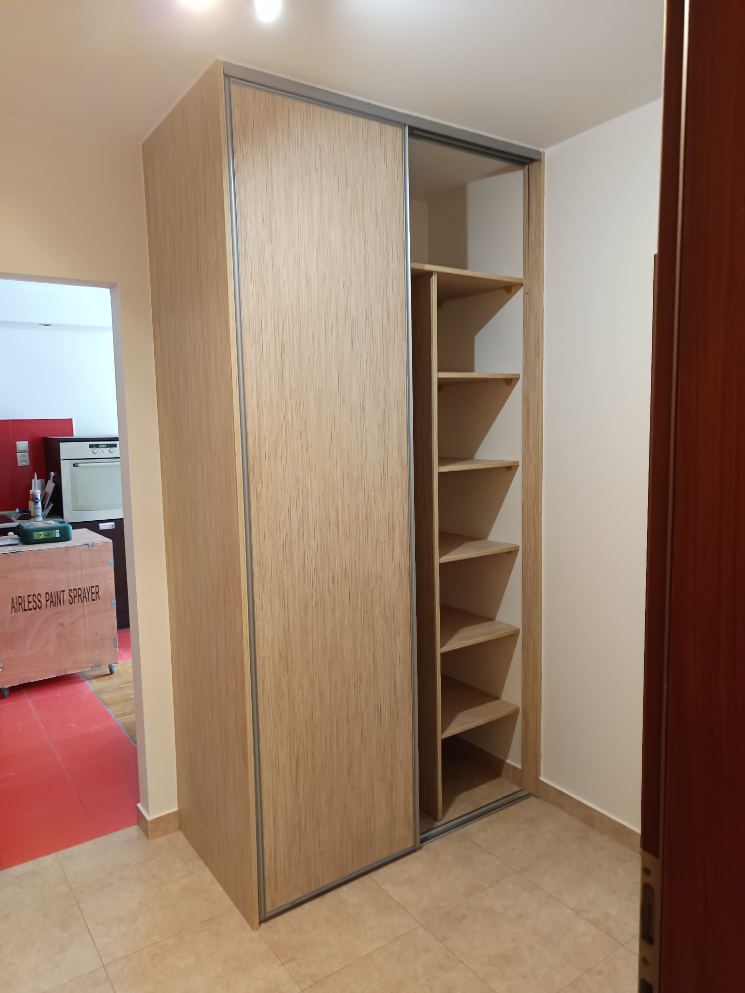 Wysoka szafa wnękowa z przesuwnymi drzwiami o teksturze jasnego drewna, z otwartym regałem z półkami po prawej stronie, widoczna w przedpokoju z płytkami podłogowymi.