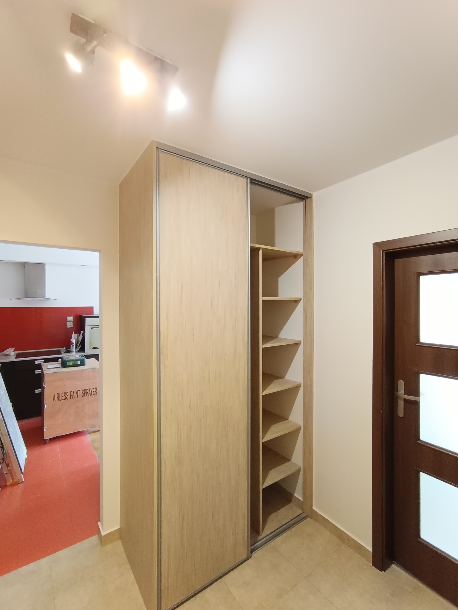 Nowoczesna szafa wnękowa z przesuwnymi drzwiami o fakturze drewna, widoczne półki wewnątrz, obok ciemne drzwi wejściowe, w tle fragment kuchni z czerwonymi panelami.