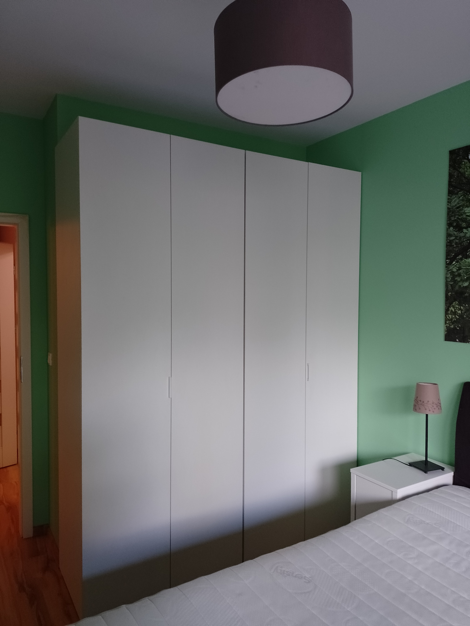 Wnętrze sypialni z zielonymi ścianami, białym, czterodrzwiowym wbudowanym szafą, lampką nocną na białej szafce i częścią łóżka z materacem.