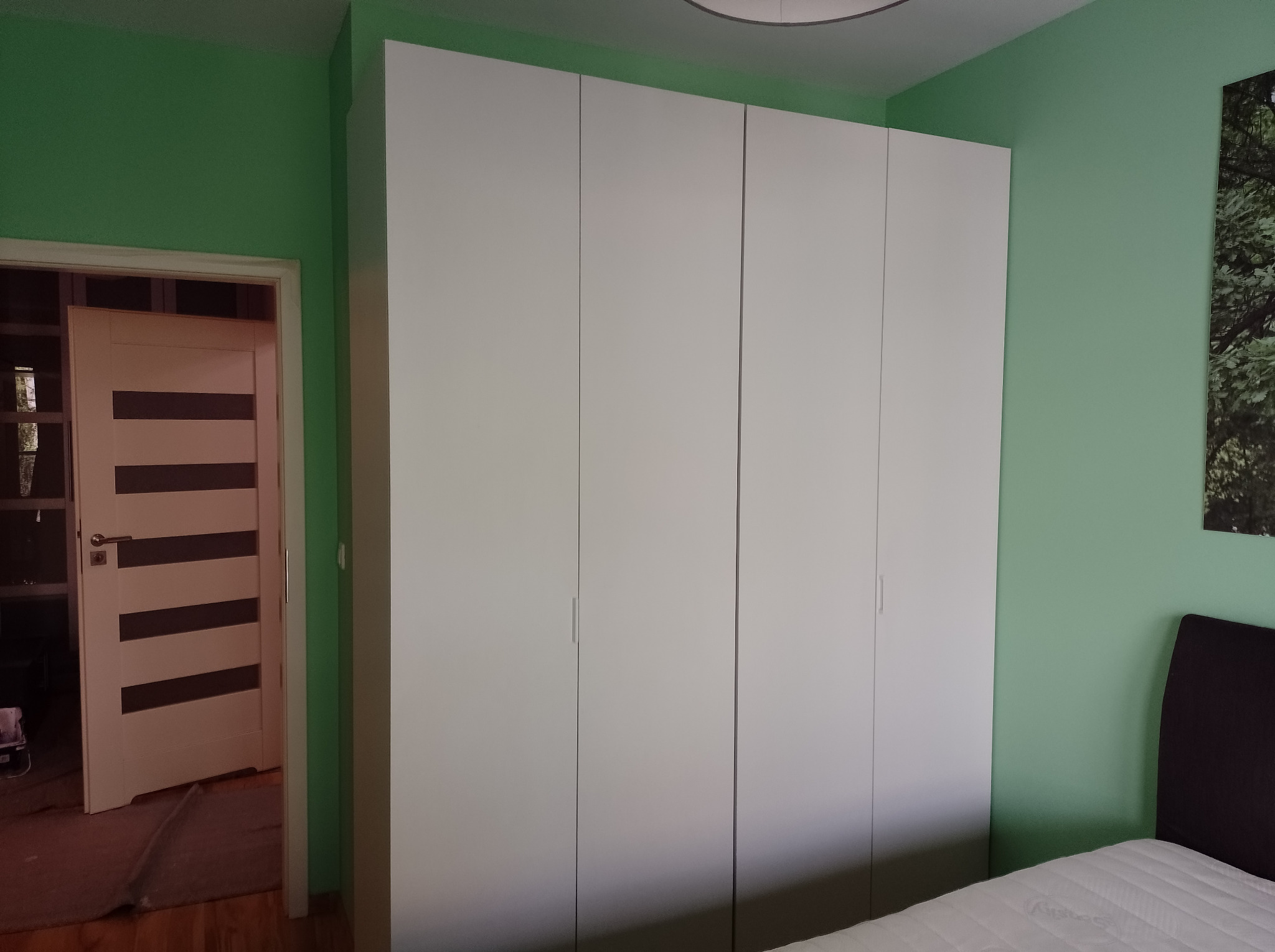 Pokój z zielonymi ścianami, białym czterodrzwiowym segmentem szafy i widocznym fragmentem łóżka z zagłówkiem oraz otwartymi drzwiami do innego pomieszczenia.