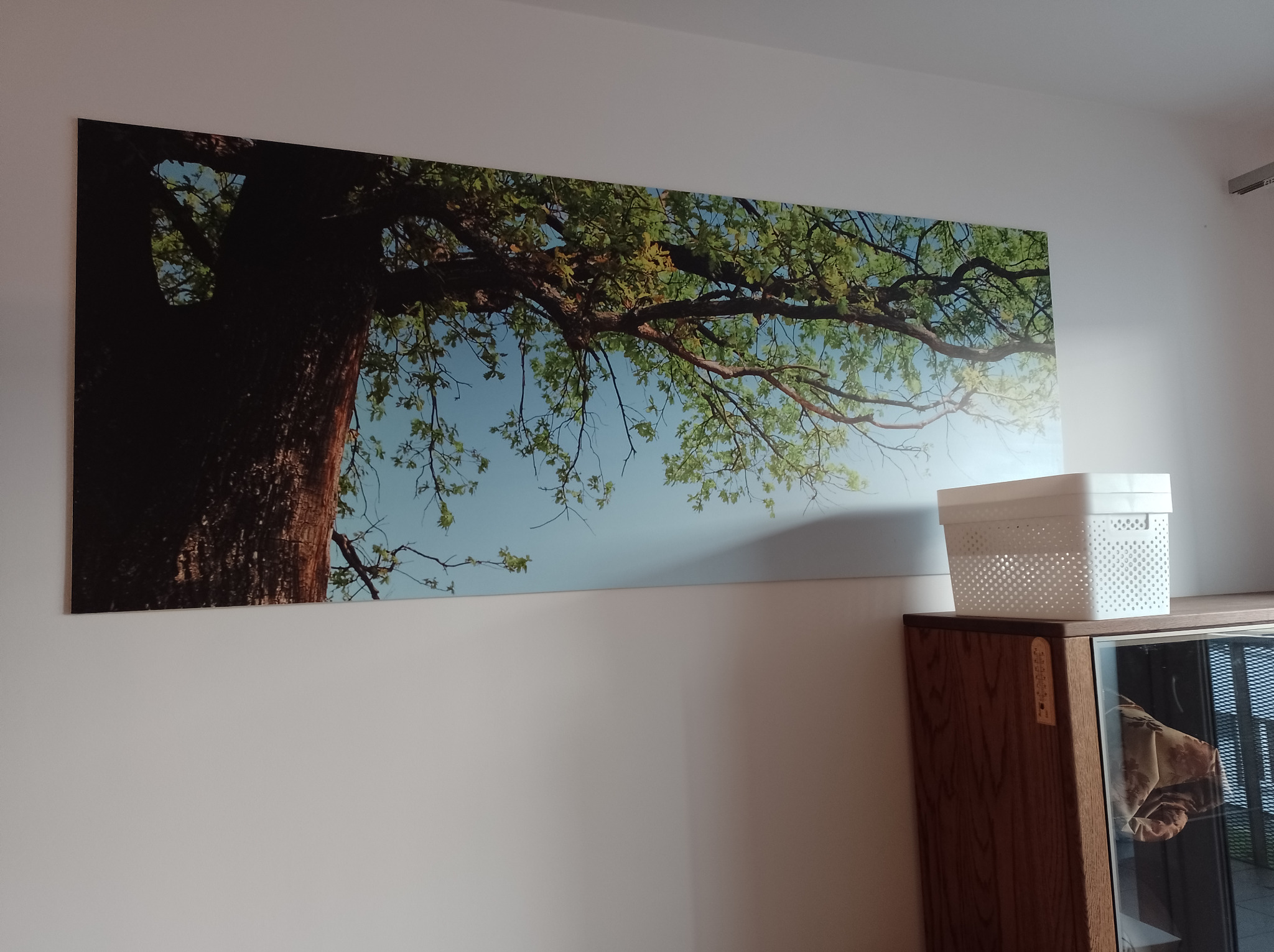 Wnętrze pokoju z długim, panoramicznym obrazem drzewa na ścianie, umieszczonym nad drewnianą komodą z przeszklonymi drzwiczkami i białym koszykiem na górze.