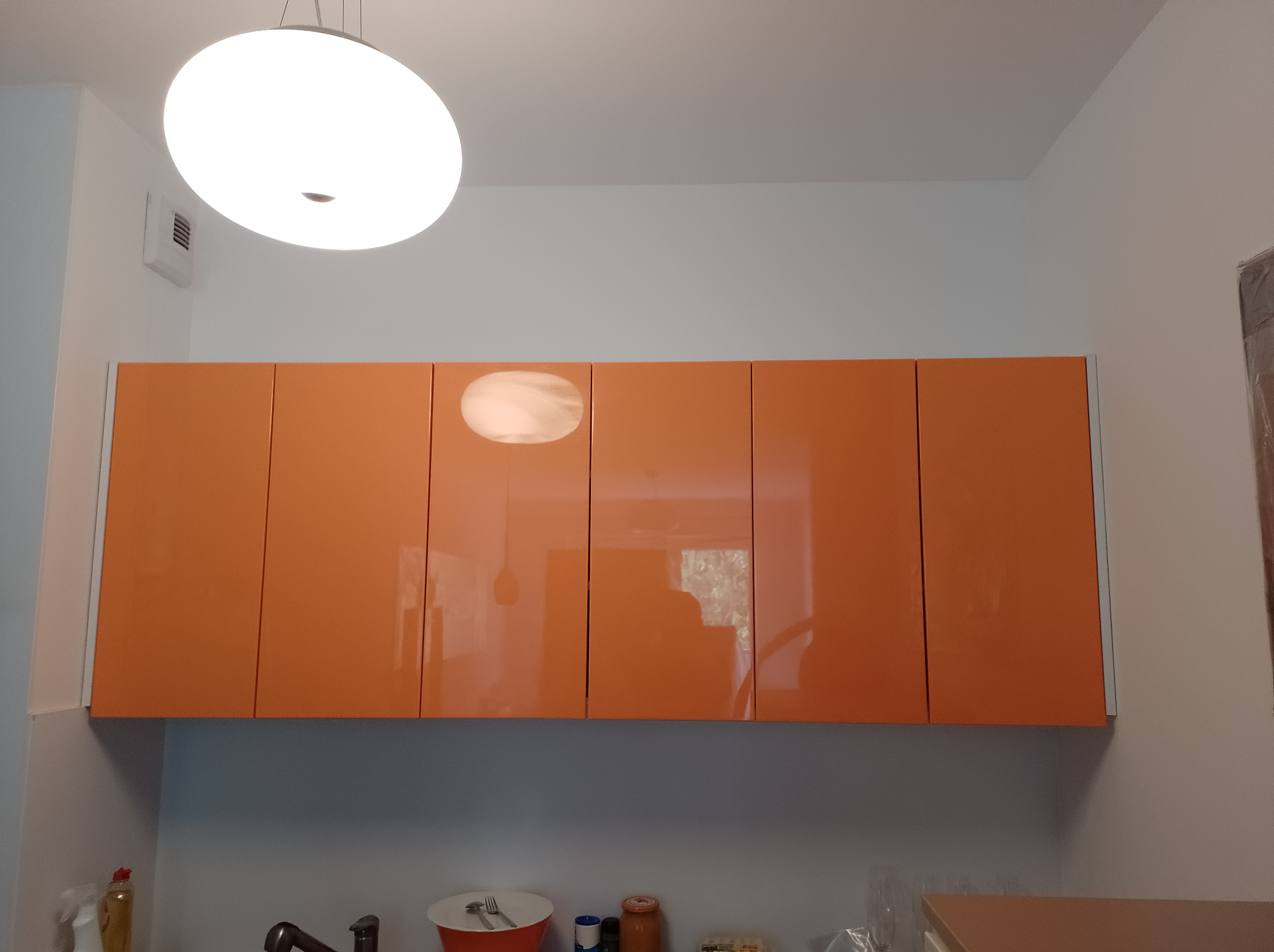Pomarańczowe szafki kuchenne wiszące z błyszczącymi frontami, oświetlone okrągłą lampą sufitową, widok od dołu.