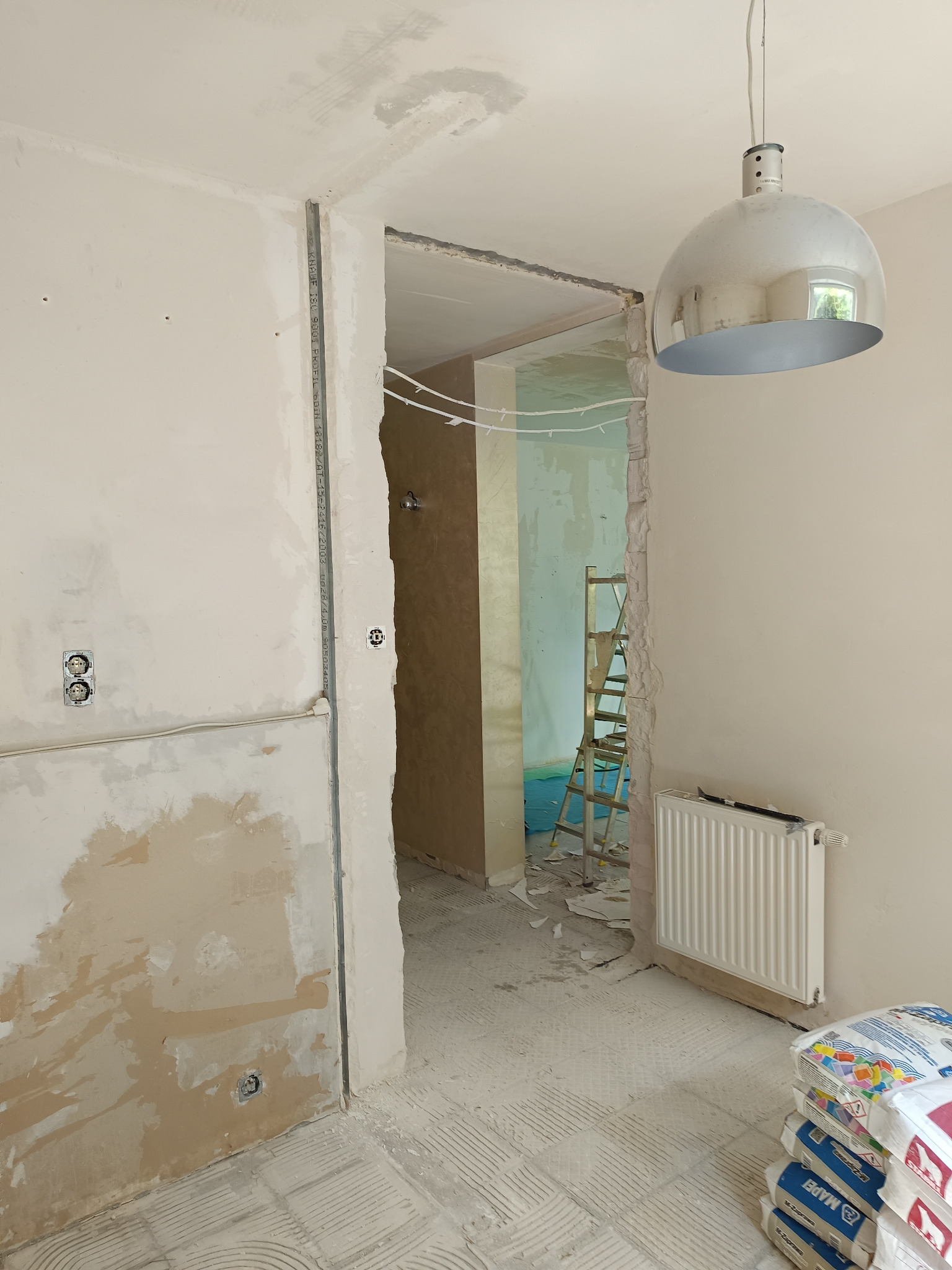 Wnętrze w trakcie remontu: ściany częściowo przygotowane do malowania, widoczne puszki elektryczne, listwa montażowa, grzejnik, drabina w tle i worki z zaprawą na podłodze.
