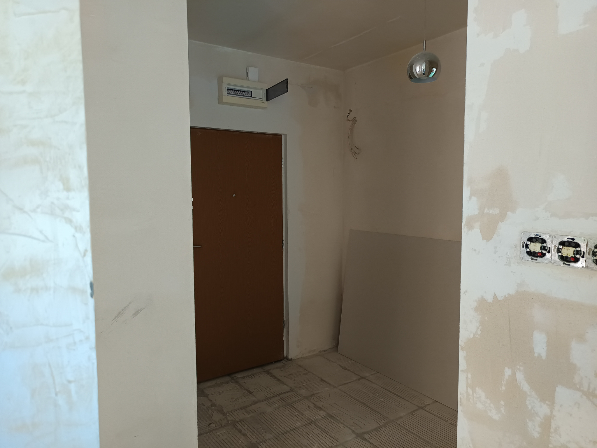 Korytarz w trakcie prac przygotowawczych: ściany z widocznymi śladami szpachlowania, niezabezpieczone gniazdka elektryczne, płyta gipsowo-kartonowa oparta o ścianę, odkryta skrzynka elektryczna...