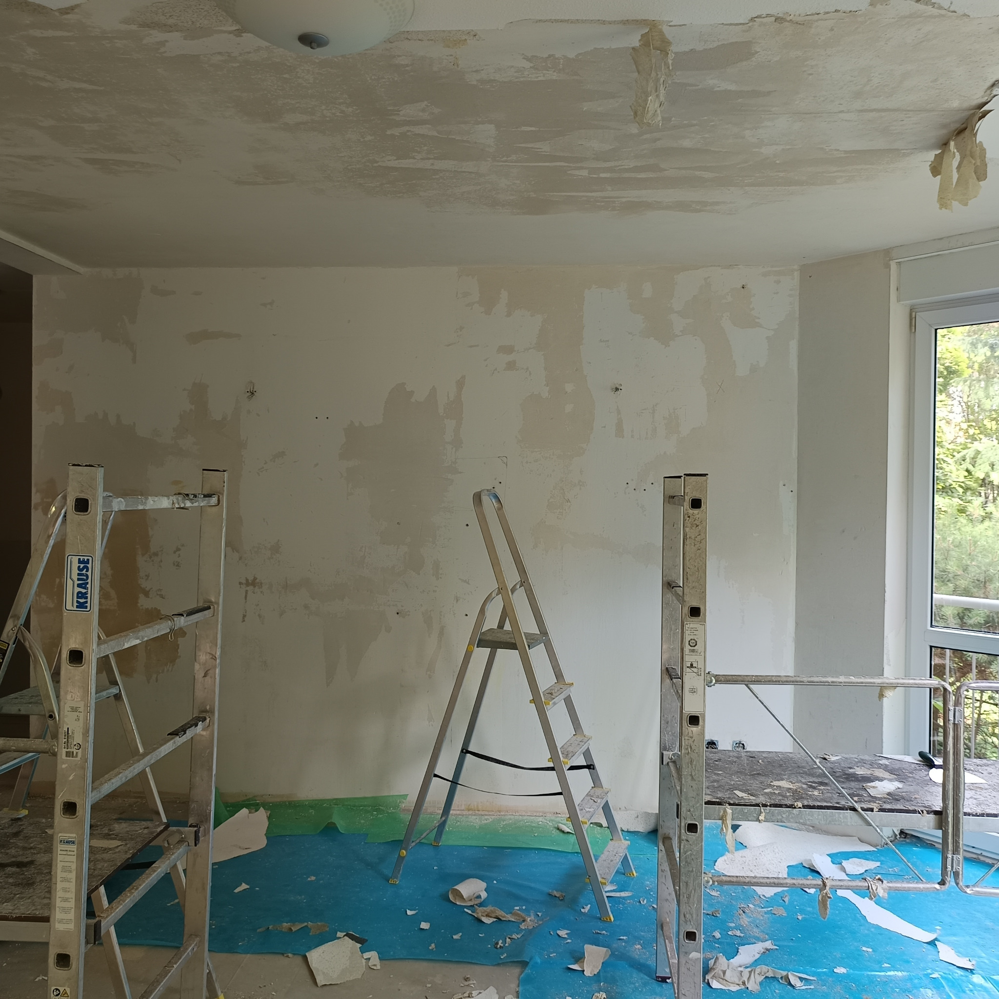 Pokój w trakcie przygotowania do prac wykończeniowych: ściany z resztkami starej farby, fragmenty odchodzącej warstwy sufitu, rozłożona folia ochronna na podłodze i rozstawione drabiny Krause.