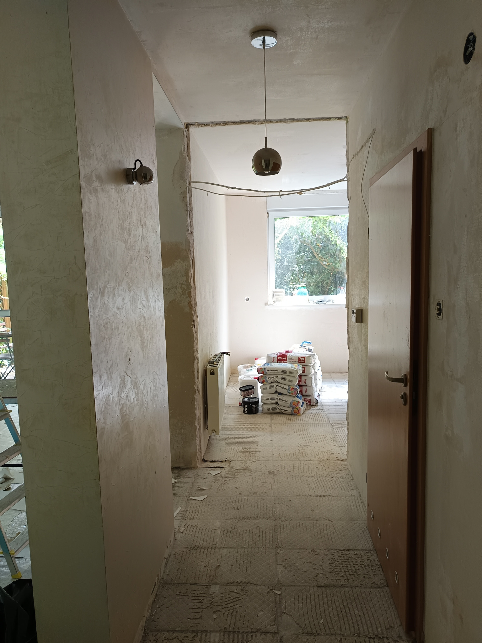 Korytarz w trakcie prac przygotowawczych do remontu: ściany częściowo pokryte fakturą, podłoga z resztkami kleju, w głębi sterta worków z zaprawą.