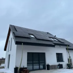 Biały dom jednorodzinny z ciemną dachówką i panelami fotowoltaicznymi na dachu w pochmurny dzień, widoczne okna dachowe i częściowo ośnieżony teren wokół.