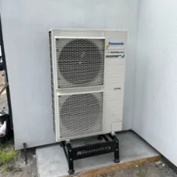 Zewnętrzna jednostka pompy ciepła Panasonic zamontowana na czarnej, metalowej podstawie z widocznym logo firmy instalacyjnej na tle szarej ściany budynku.