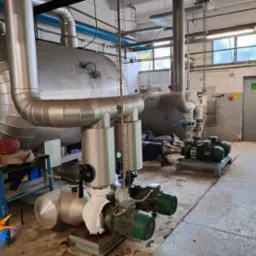 Duże, izolowane zbiorniki i pompy w pomieszczeniu technicznym, widoczne rury i drabina.