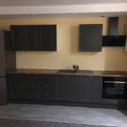 Kompletna, szara zabudowa kuchenna z drewnianym blatem, srebrną lodówką i czarnym okapem na tle żółtej ściany.