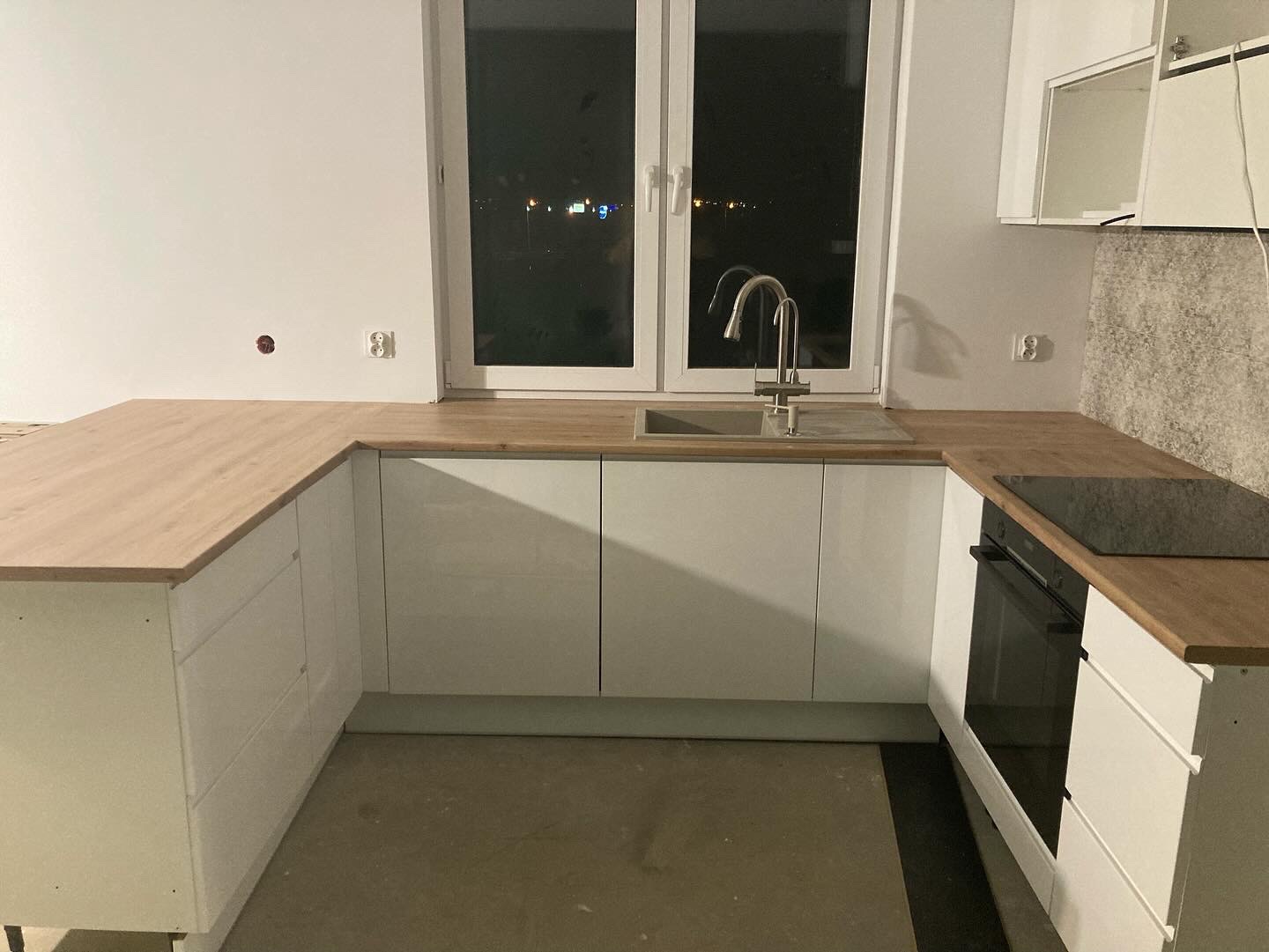 Wnętrze kuchni w trakcie montażu, widoczne białe szafki z drewnianym blatem, okno z widokiem na nocne miasto i niedokończone wykończenie ściany.