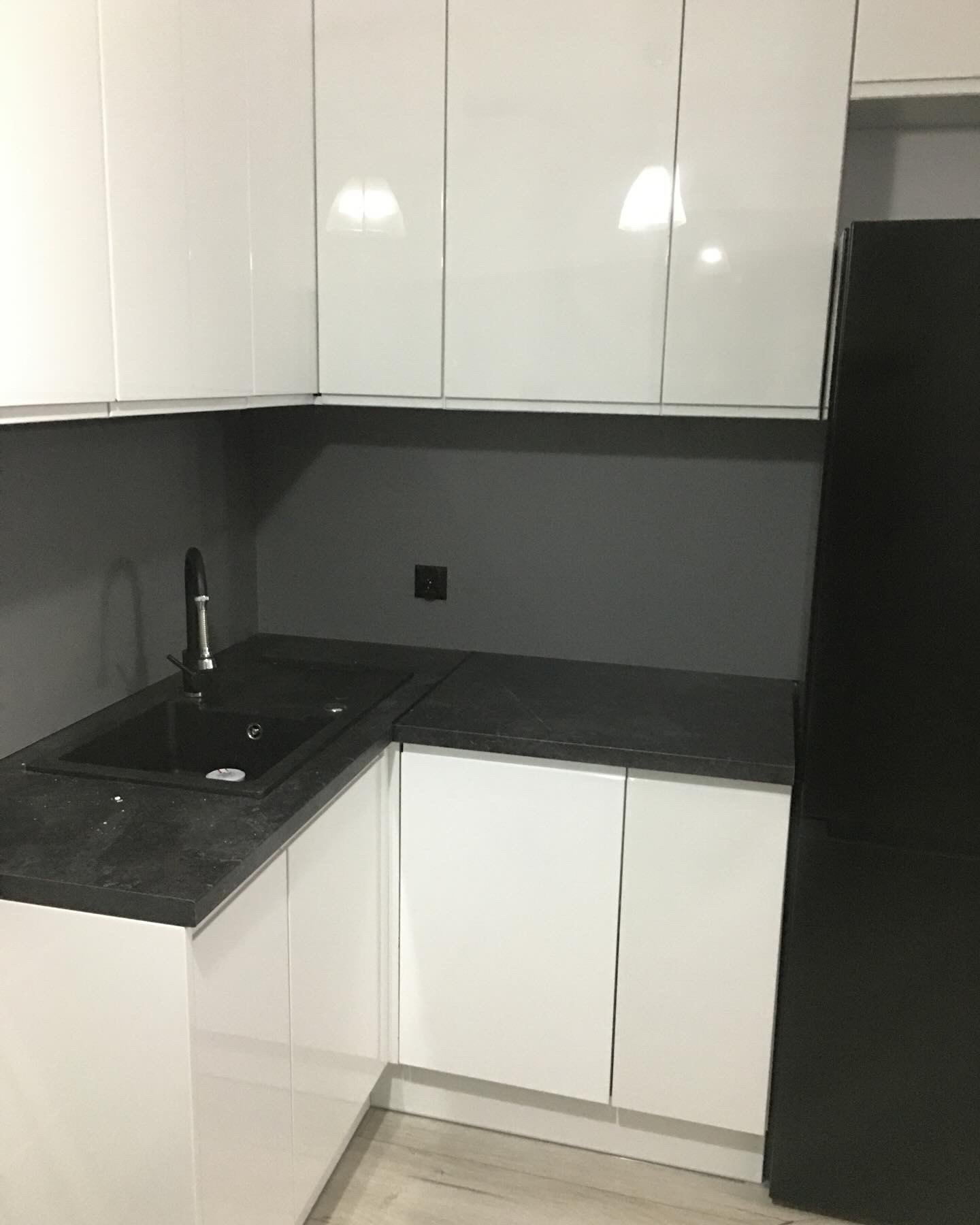 Nowoczesna kuchnia z białymi szafkami na wysoki połysk, czarnym blatem i zlewozmywakiem, szara ściana nad blatem, widoczna czarna lodówka po prawej stronie.