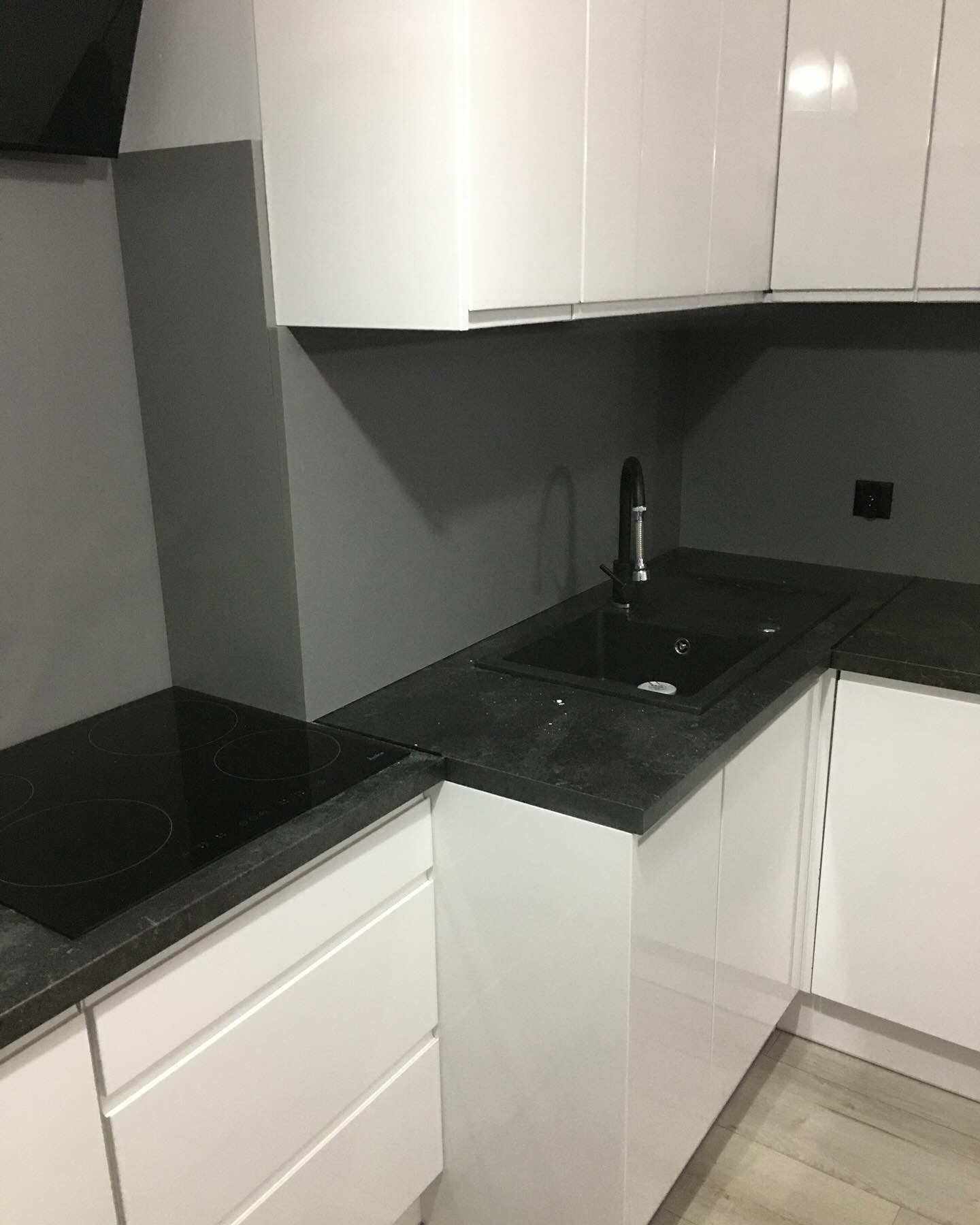 Nowoczesna, minimalistyczna kuchnia: białe, lakierowane szafki z czarnym blatem i płytą indukcyjną, czarny zlew z chromowaną baterią na tle szarej ściany.