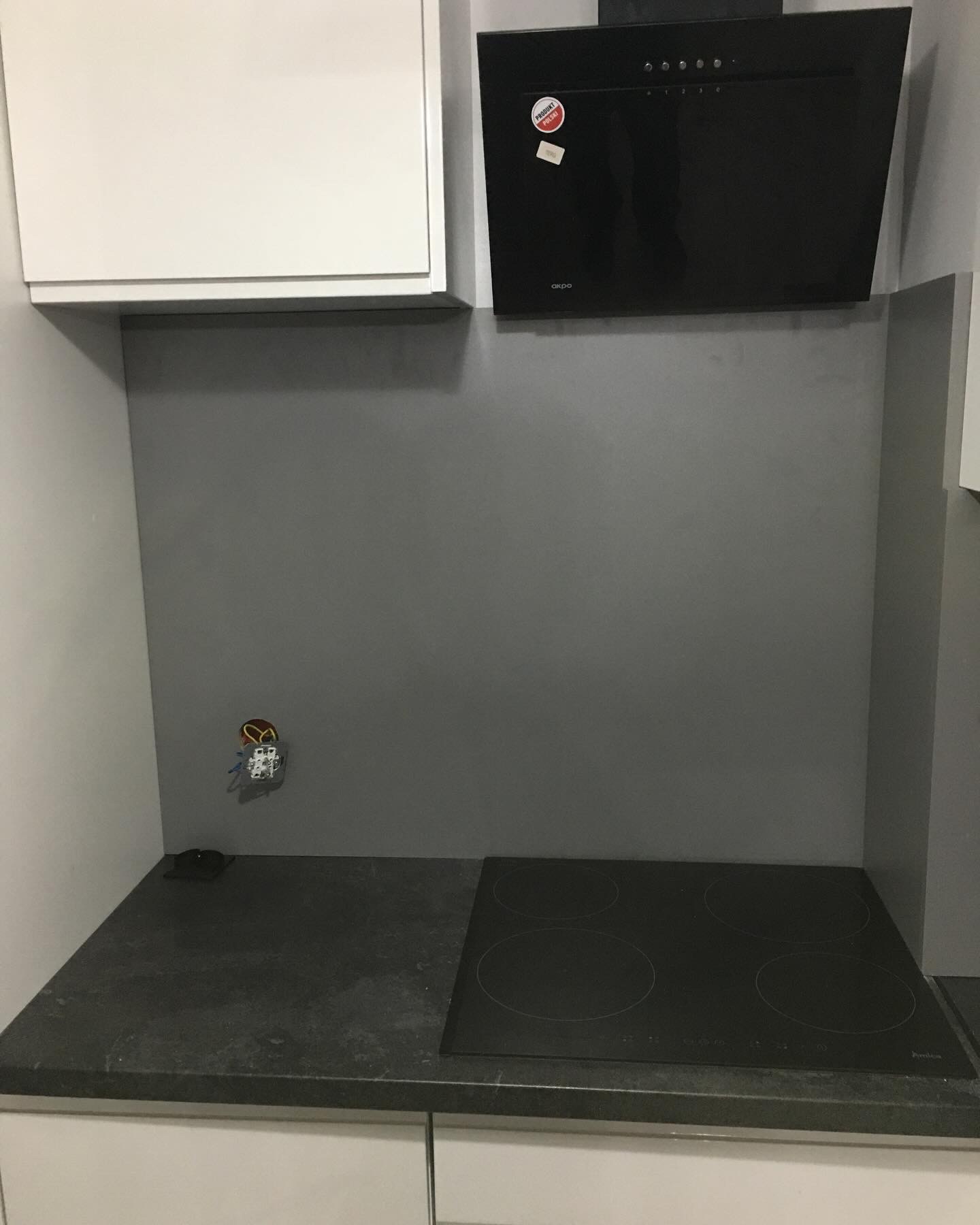 Fragment kuchni z czarną płytą indukcyjną, szarym panelem ściennym, białymi szafkami i czarnym okapem z naklejką 'Produkt Polski', widoczne gniazdko elektryczne z wystającymi kablami.