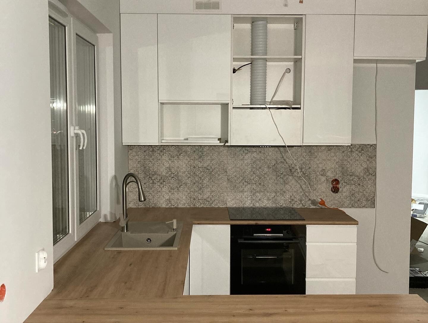 Fragment montażu białej kuchni z drewnianym blatem, widoczny zlew przy oknie, płyta indukcyjna i piekarnik; w szafce nad płytą widoczna rura wentylacyjna.