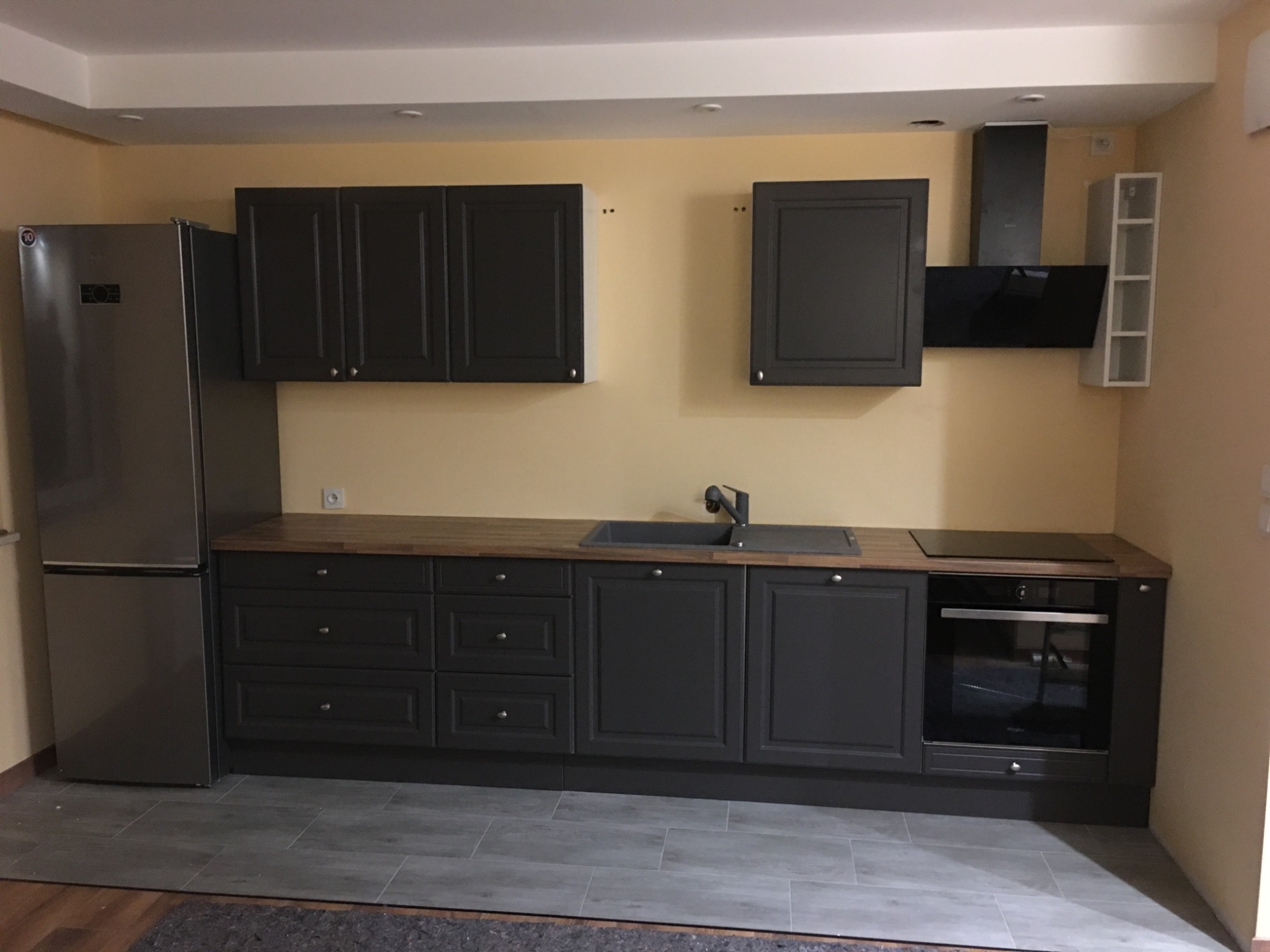 Kompletna, szara zabudowa kuchenna z drewnianym blatem, srebrną lodówką i czarnym okapem na tle żółtej ściany.