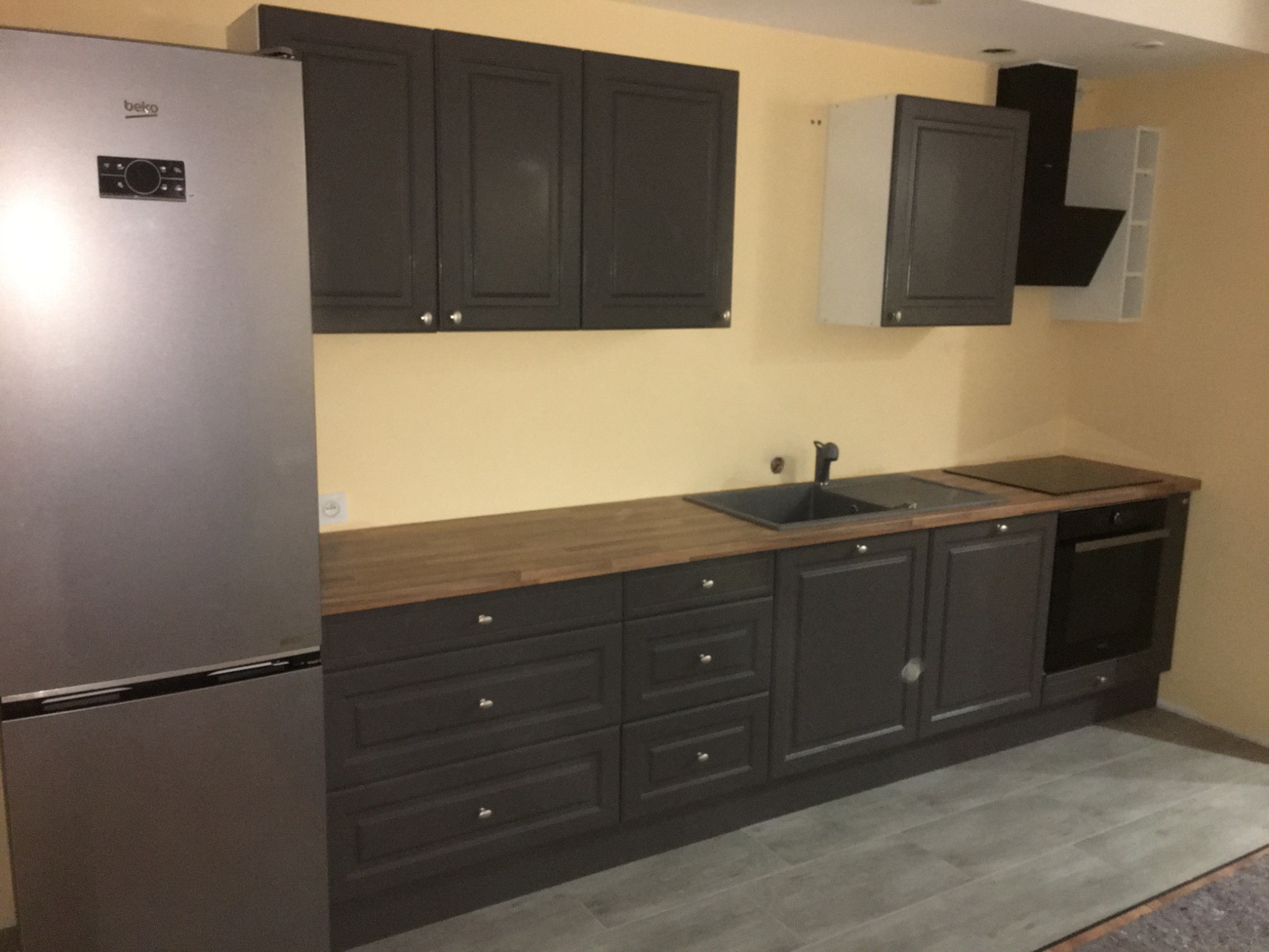 Nowoczesna szara kuchnia z drewnianym blatem, srebrną lodówką Beko i czarnym okapem, widziana pod kątem z szarymi panelami podłogowymi.