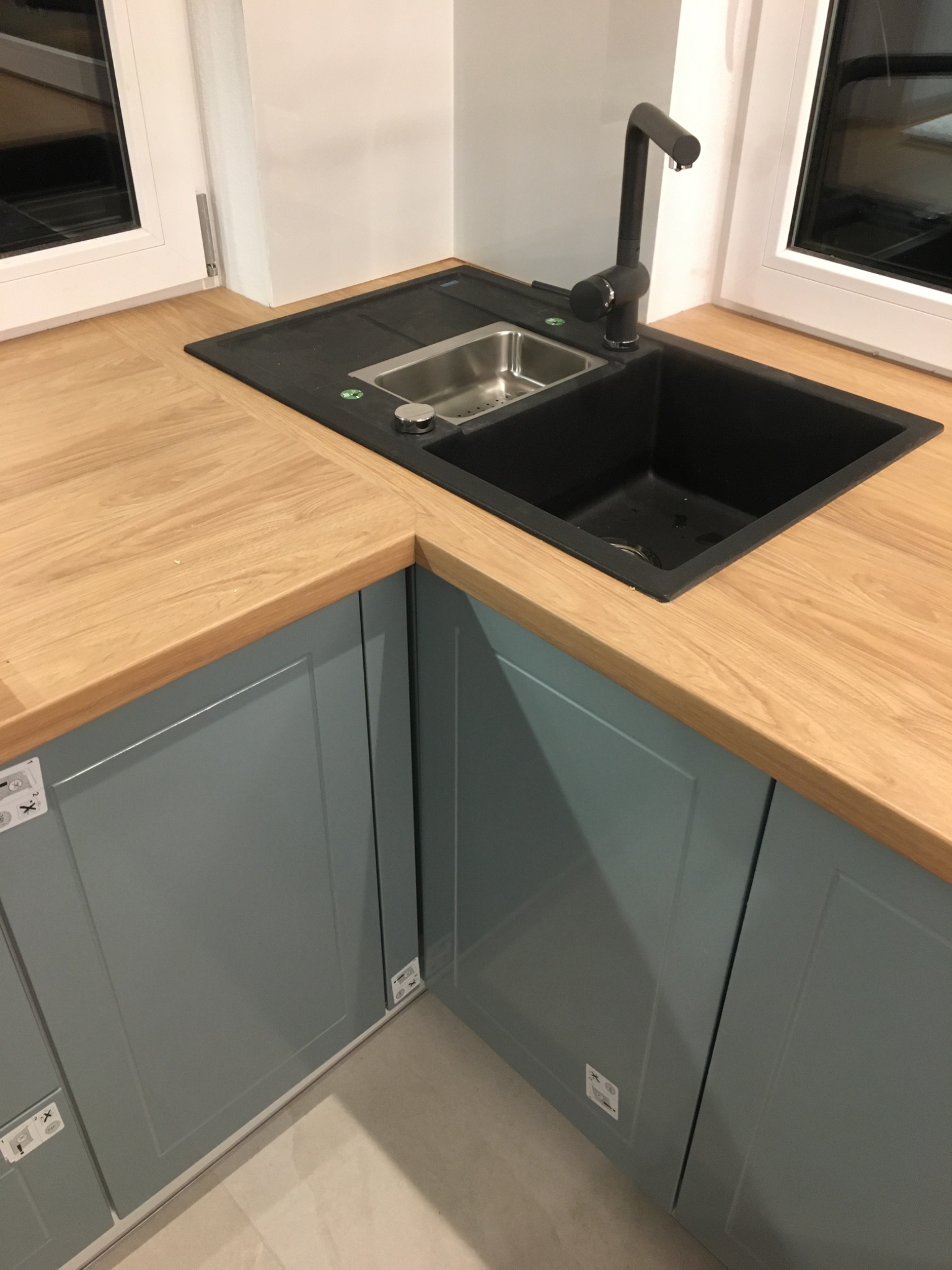Fragment kuchni z drewnianym blatem, czarnym zlewem i szarozielonymi szafkami z panelowymi frontami, widoczne okno po lewej i prawej stronie zlewu.